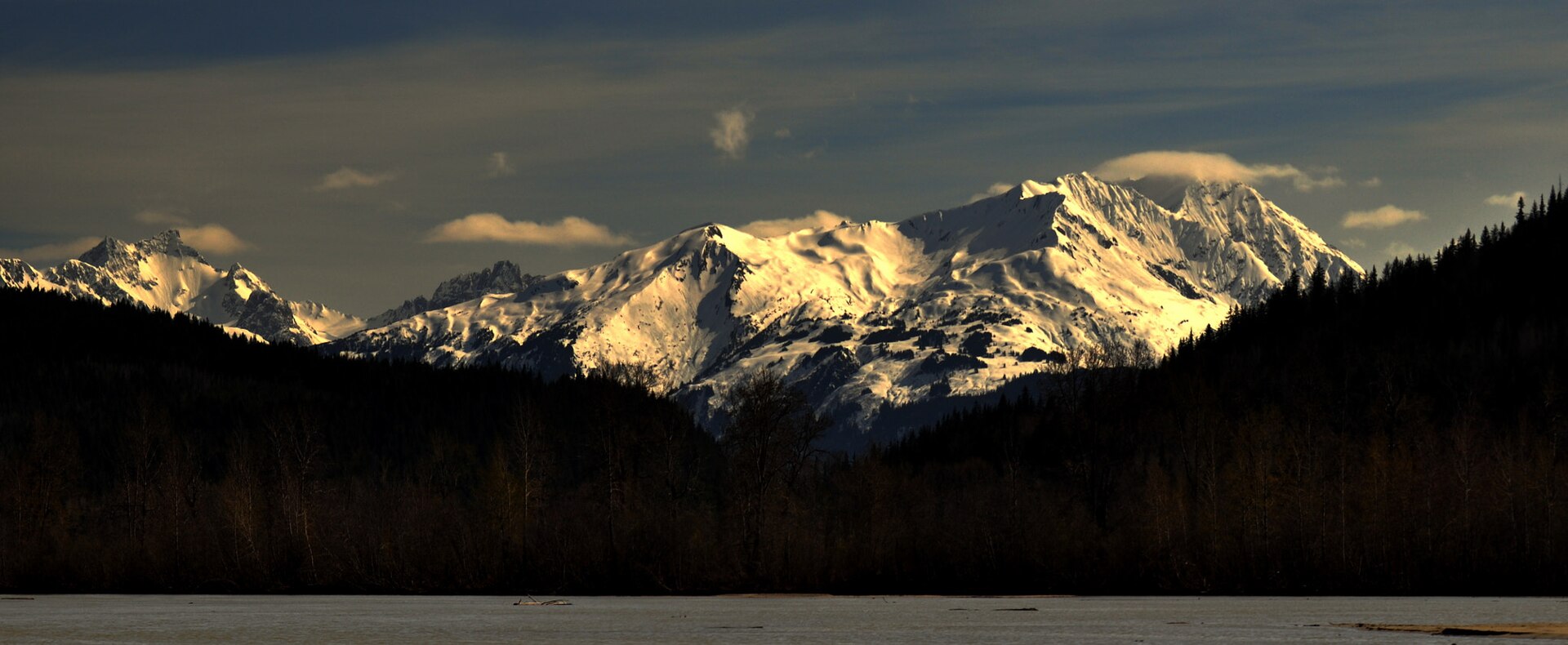 Sunlit snow-covered mountains above dark forest in Southeast Alaska