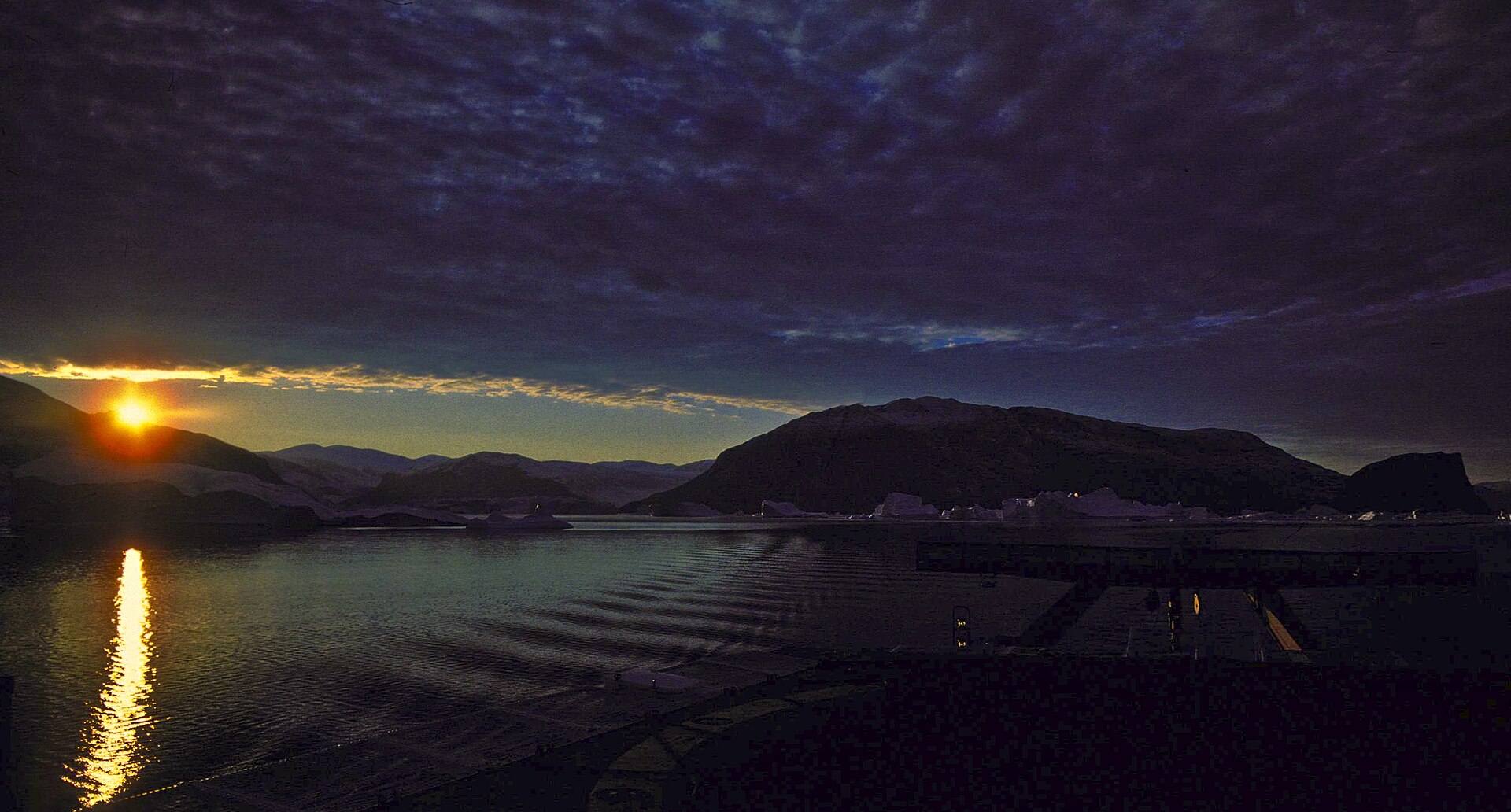 Golden sunset reflected across the still water and dark mountains of Scoresby Sound