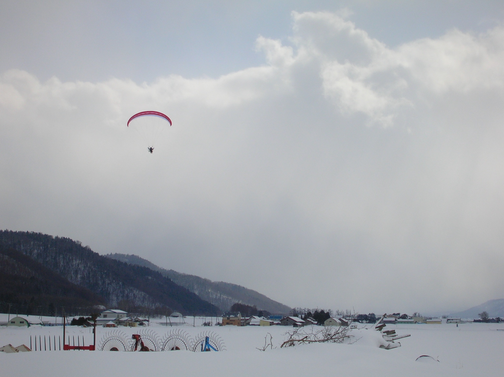 A paraglider crossing a pale winter sky above snowy Furano fields