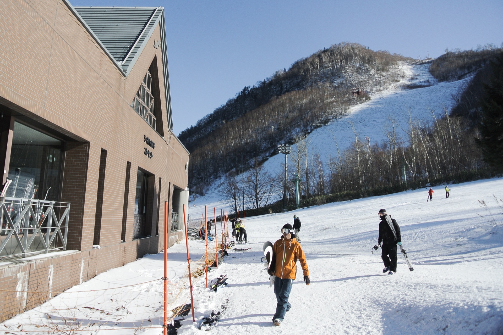 Skiers and snowboarders outside the base lodge at Sapporo Teine, with a pisted run and chairlift behind