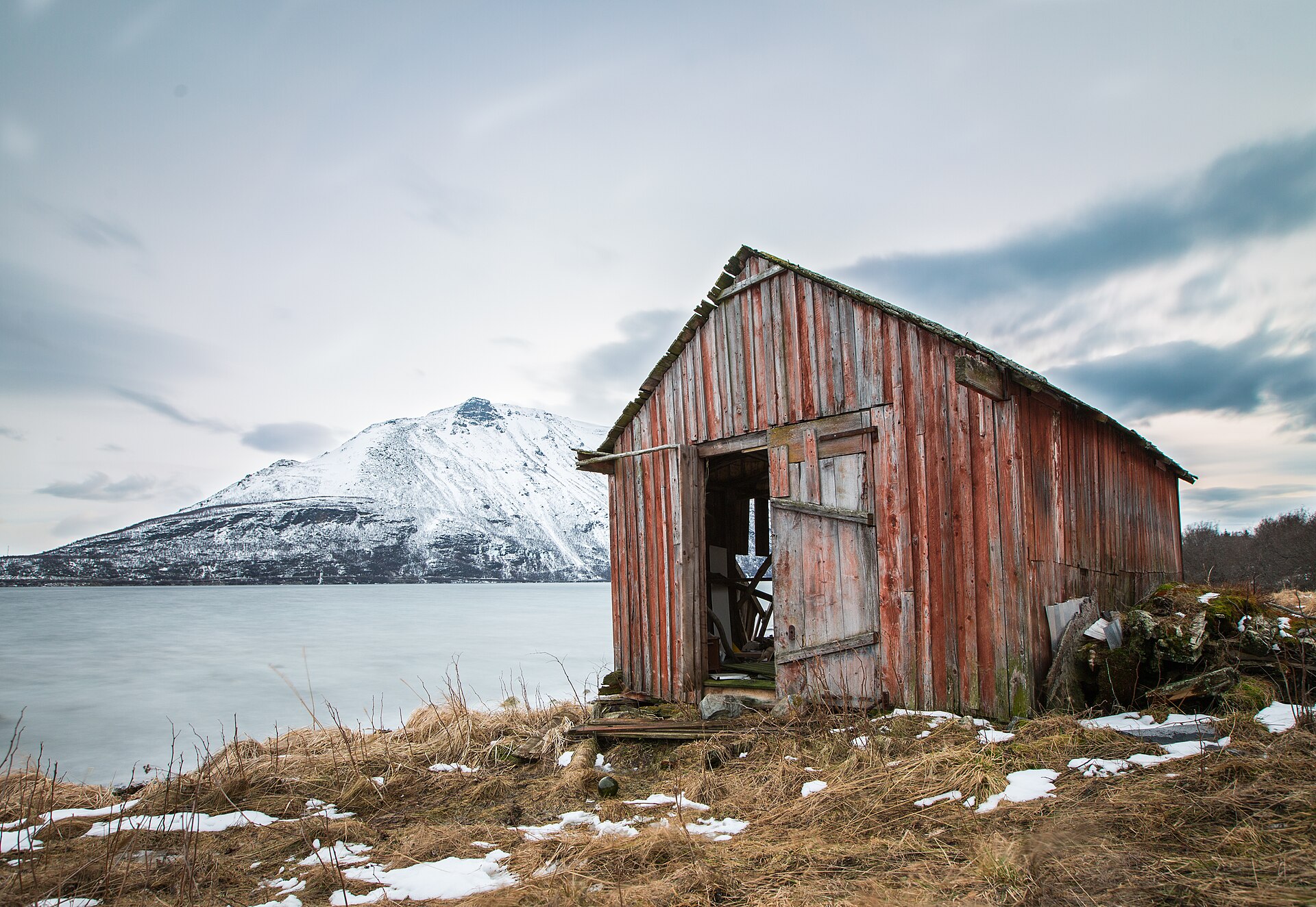 A weathered red timber building beside the fjord beneath a snowy Lyngen mountain