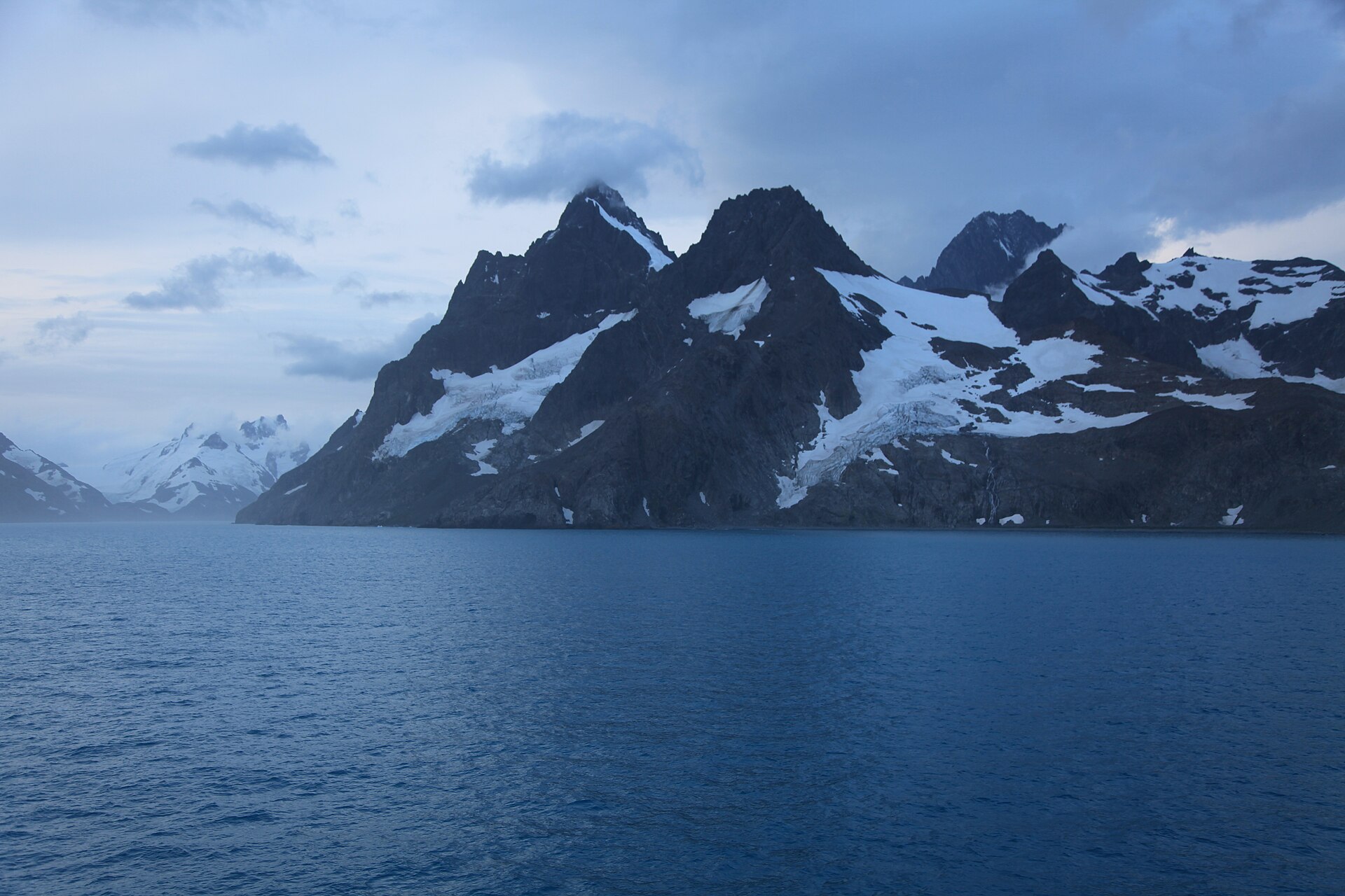 Black alpine peaks and glaciers lining Drygalski Fjord under a pale sky