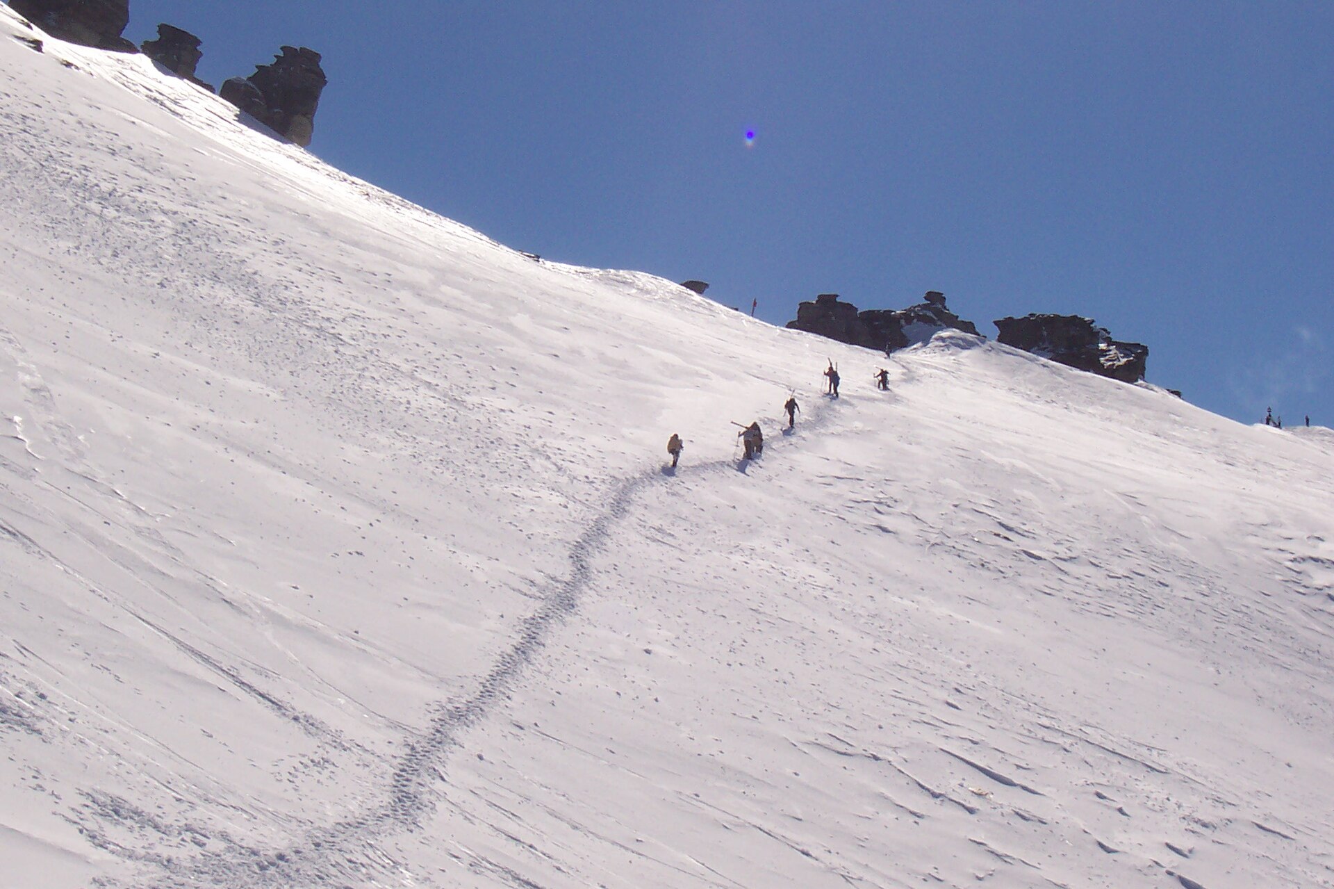 A group climbing a broad snow slope toward the summit of Treble Cone