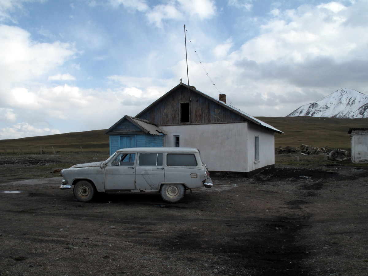 A simple roadside building and vehicle beneath the mountains of Suusamyr