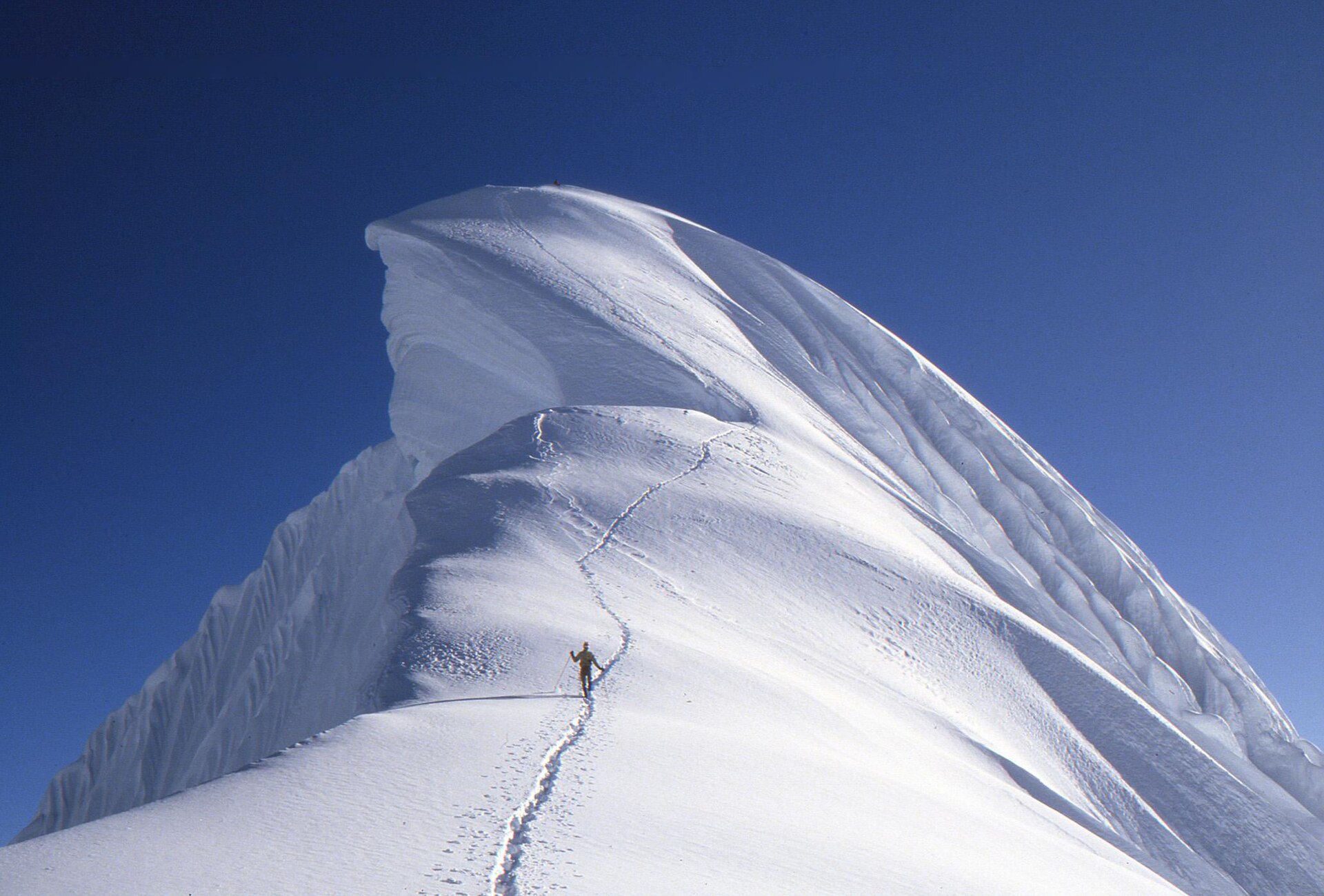 A mountaineer climbing a narrow sculpted snow ridge in the Cordillera Blanca